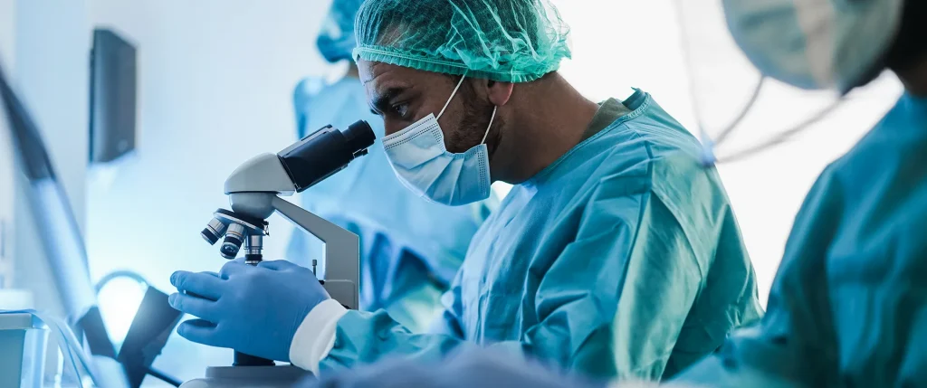 Médico con mascarilla analizando muestras en un laboratorio a través de un microscopio junto a otros profesionales sanitarios